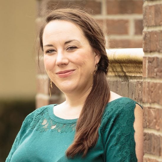 woman with dark brown hair, green shirt, smiling at the camera and leaning against a brick wall